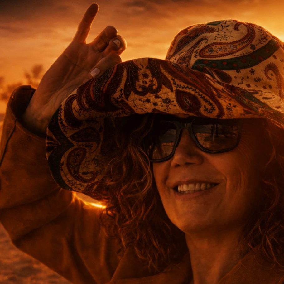 Mujer sonriente con gafas y sombrero colorido, levantando la mano al atardecer.