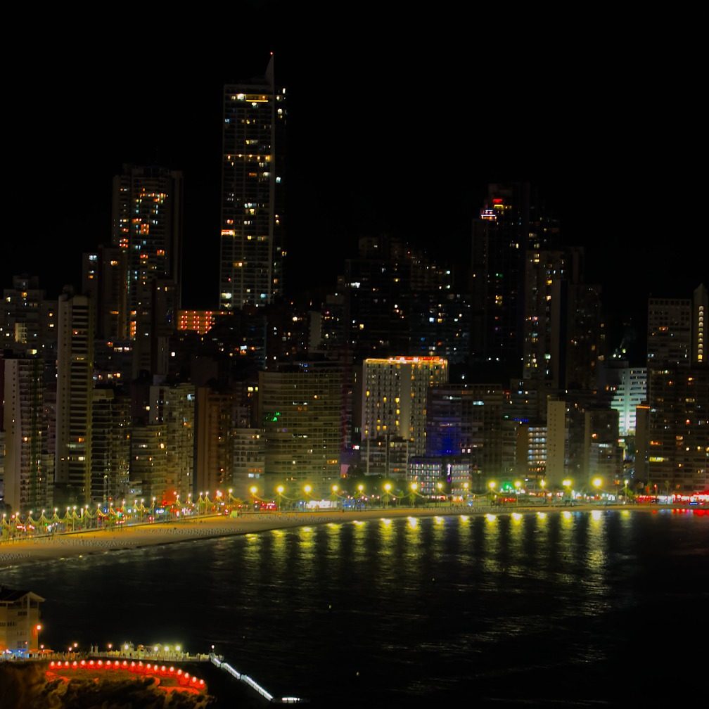 Vista nocturna de Benidorm que es una ciudad costera de la comunidad valenciana, iluminada, con rascacielos y el mar reflejando luces.
